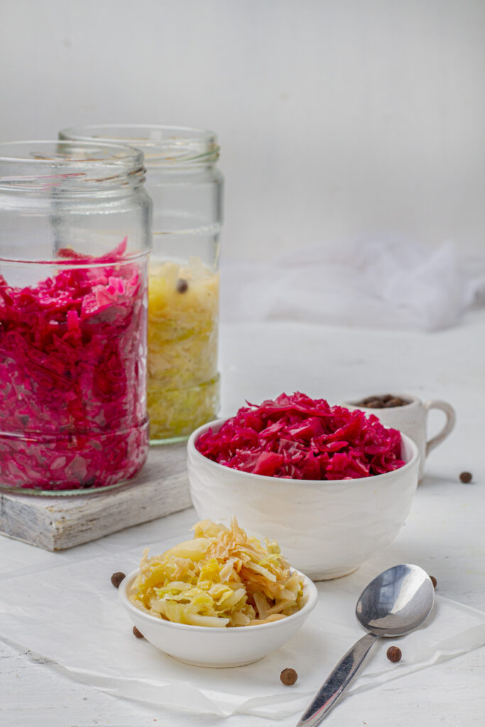  Jars of homemade sauerkraut made from white and purple cabbage, along with a small bowl of purple cabbage sauerkraut, all displayed on a white background.