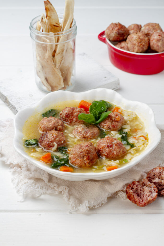 Italian wedding soup, served in a white bowl, surrounded with bread and sausage meatballs.