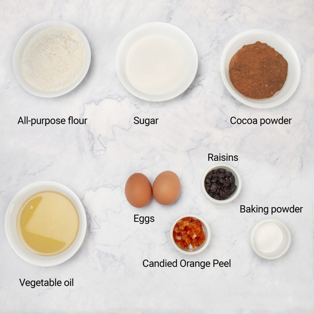 Ingredients for Christmas Chocolate Muffins, including flour, cocoa powder, sugar, eggs, oil, baking powder, candied orange peel, and raisins, arranged on a countertop.