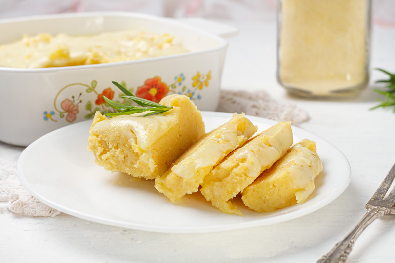 A golden-brown slice of creamy polenta, topped with a layer of melted cheese, is served on a white plate. A casserole dish filled with additional polenta can be seen in the background.