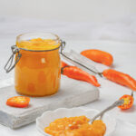 A glass jar filled with vibrant orange-yellow aji amarillo paste, a Peruvian condiment, sits beside fresh aji amarillo peppers on a wooden cutting board.