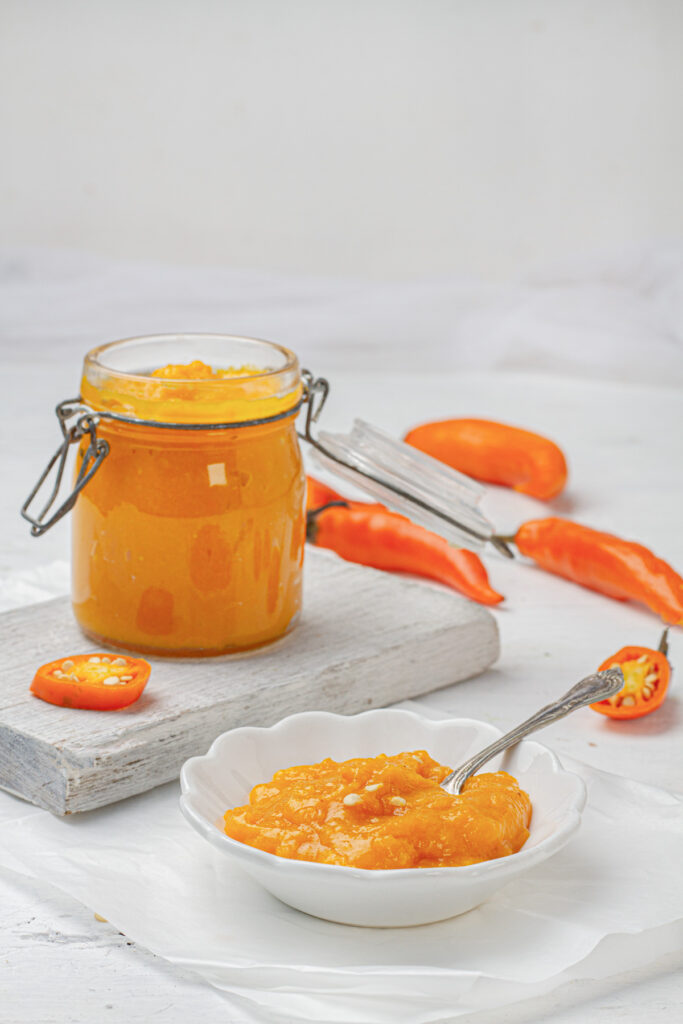 A glass jar filled with vibrant orange-yellow aji amarillo paste, a Peruvian condiment, sits beside fresh aji amarillo peppers on a wooden cutting board.