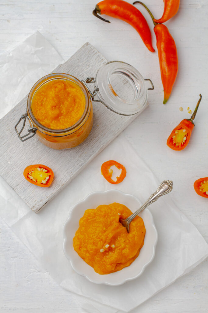 Top view a glass jar filled with yellow aji amarillo paste, a Peruvian condiment, sits beside fresh aji amarillo peppers sliced on a light wooden cutting board.