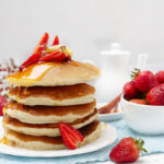 A stack of golden brown pancakes topped with fresh strawberries, butter, and a drizzle of maple syrup.