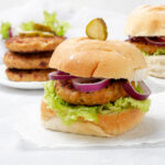 A plate of homemade chicken burgers with lettuce, tomato, onion, and pickles on a white background.