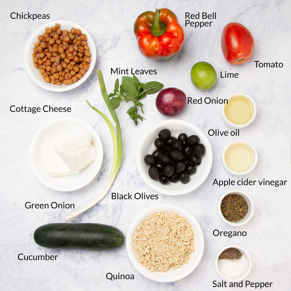 A flat lay of ingredients for a quinoa salad, including chickpeas, red bell pepper, tomato, lime, red onion, olive oil, apple cider vinegar, oregano, salt, pepper, cottage cheese, green onion, cucumber, black olives, mint, and quinoa.