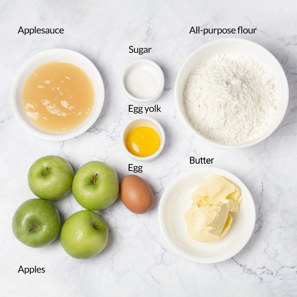 A flat lay of ingredients for making an apple pie, including apples, flour, sugar, butter, eggs, and applesauce.