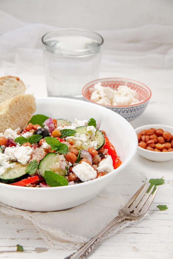 A bowl of quinoa salad topped with feta cheese, chickpeas, cucumber, red bell pepper, olives, and fresh mint.