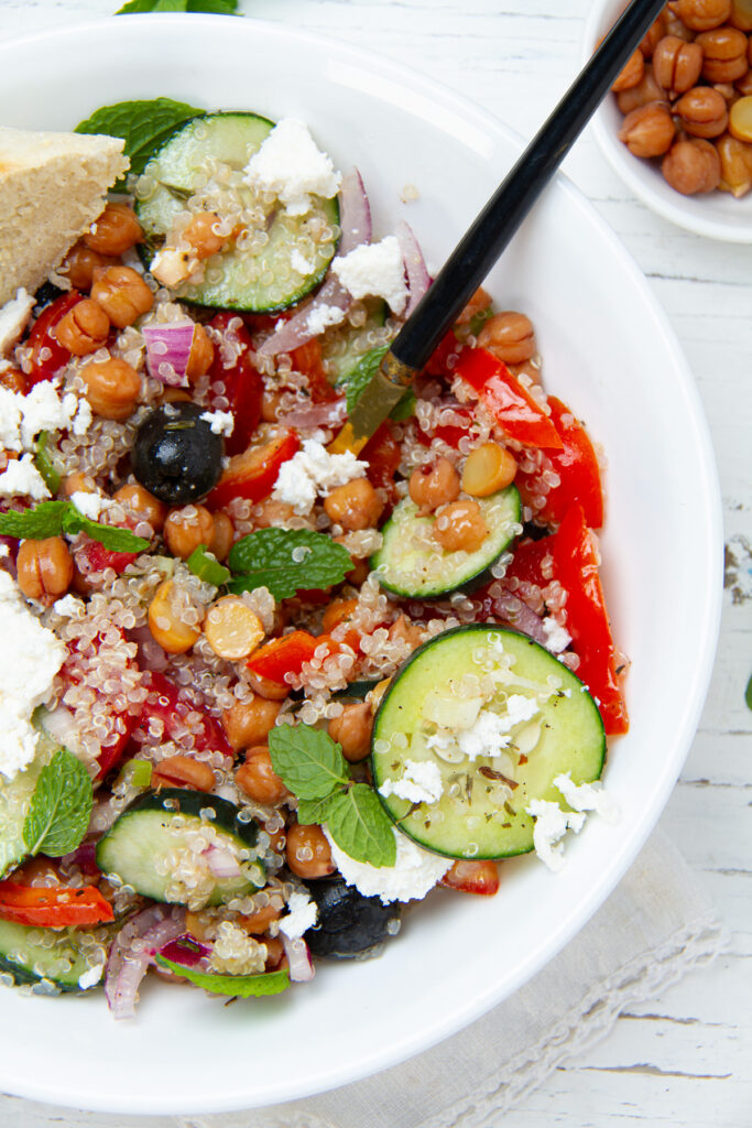 A bowl of quinoa salad topped with feta cheese, chickpeas, cucumber, red bell pepper, olives, and fresh mint.