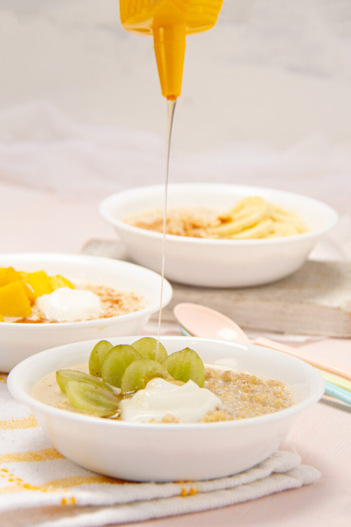 Three bowls of quinoa oatmeal with different toppings: sliced banana and cinnamon, diced mango and yogurt, and green grapes and yogurt. A jar of honey, a teapot, and a cup are also visible on a light pink wooden table.