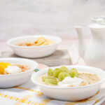 Three bowls of quinoa oatmeal with different toppings: sliced banana and cinnamon, diced mango and yogurt, and green grapes and yogurt. A jar of honey, a teapot, and a cup are also visible on a light pink wooden table.