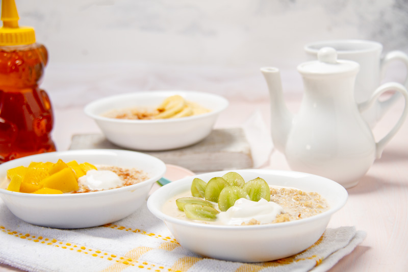 Three bowls of quinoa oatmeal with different toppings: sliced banana and cinnamon, diced mango and yogurt, and green grapes and yogurt. A jar of honey, a teapot, and a cup are also visible on a light pink wooden table.