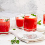 Creamy yogurt jello in three glasses, with a spoonful of jello being lifted from one, garnished with mint leaves and a small bowl of white cream.
